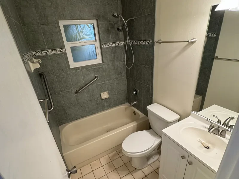 A bathroom with a beige tub against green wall tile, a window, and a white vanity.