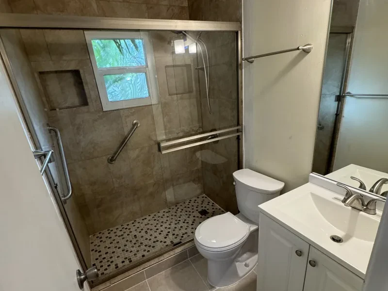 The same bathroom with a tan stone-look walk-in shower, a glass door, and the same vanity.