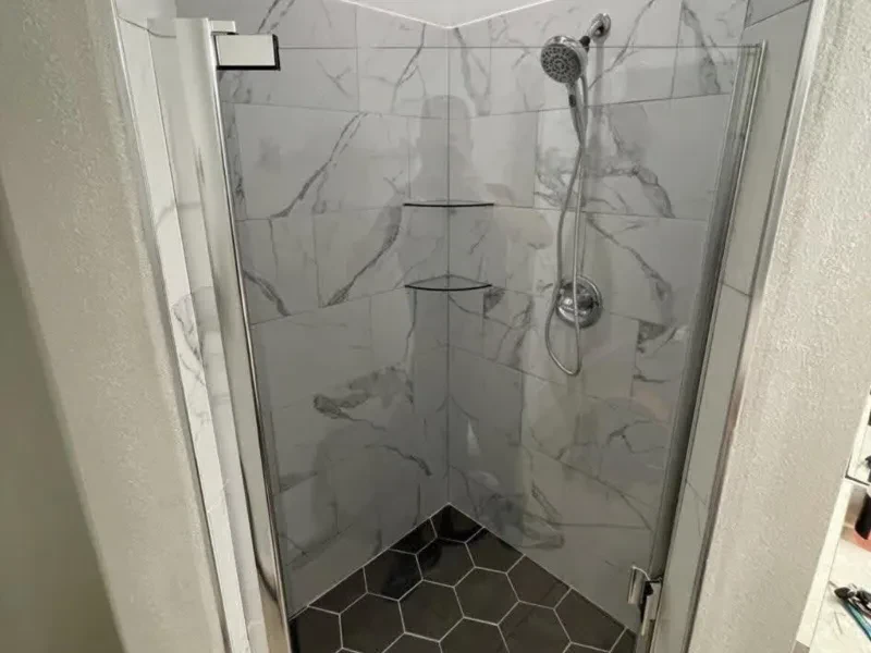 The same shower corner in gray marble-look tile, with two corner shelves and a glass panel.