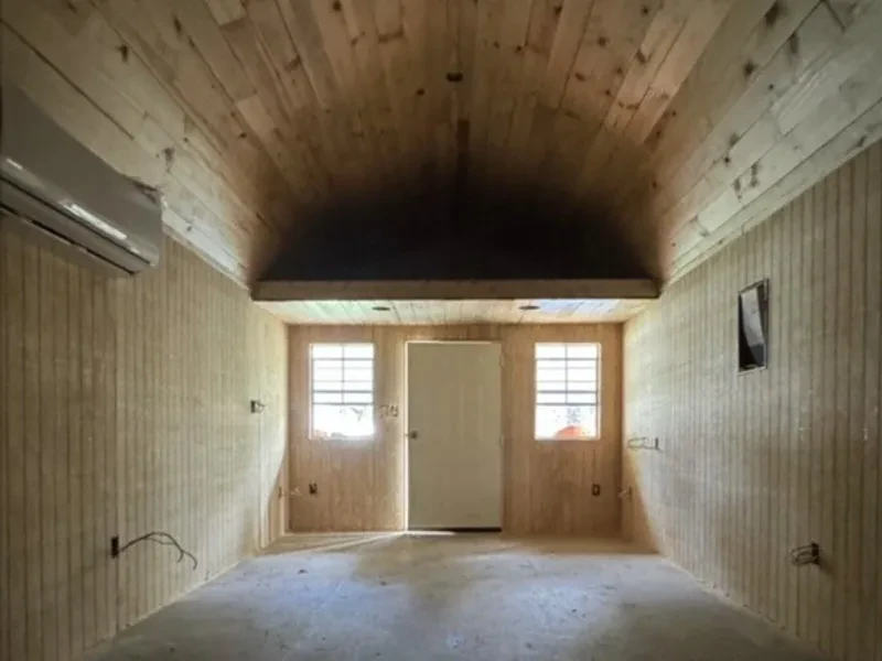 A wood-plank shed with a concrete floor, a door between two small windows, and a wall-mounted air conditioner.