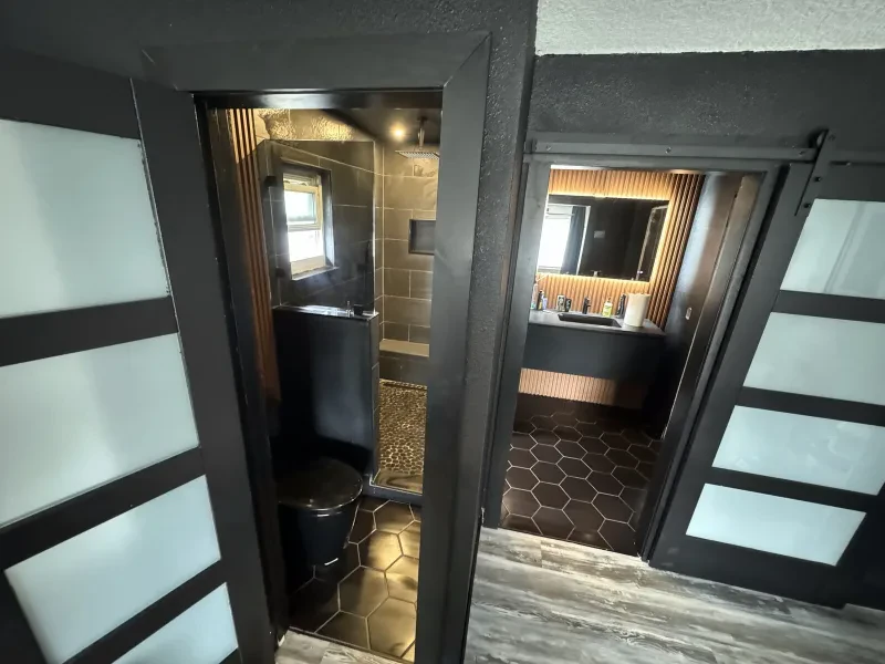 A black-tile bathroom seen through two dark doorways, with a shower on the left and a backlit mirror on the right.