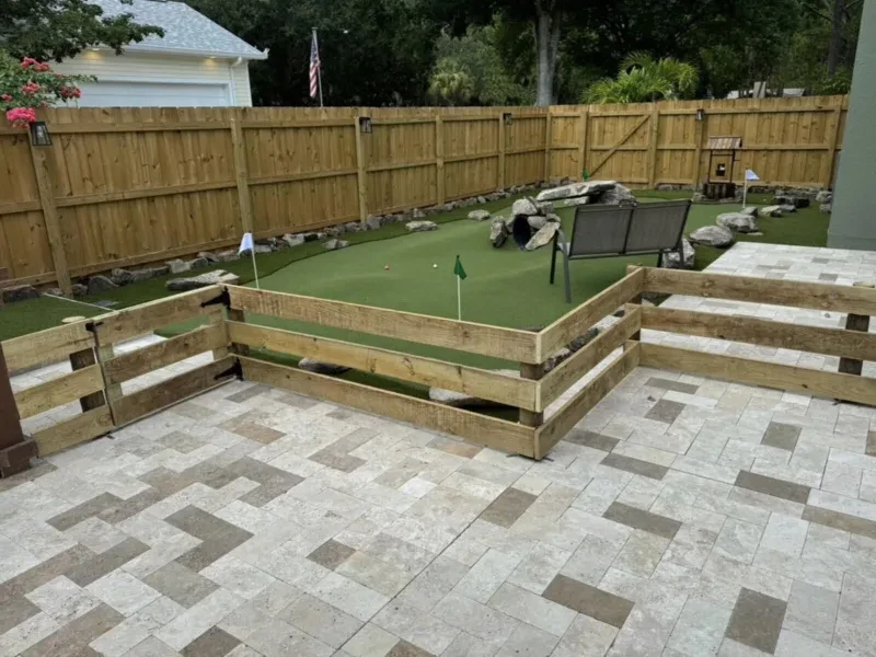 A green artificial-turf putting green beside a light paver patio, with a wood rail in front and a wood fence behind.
