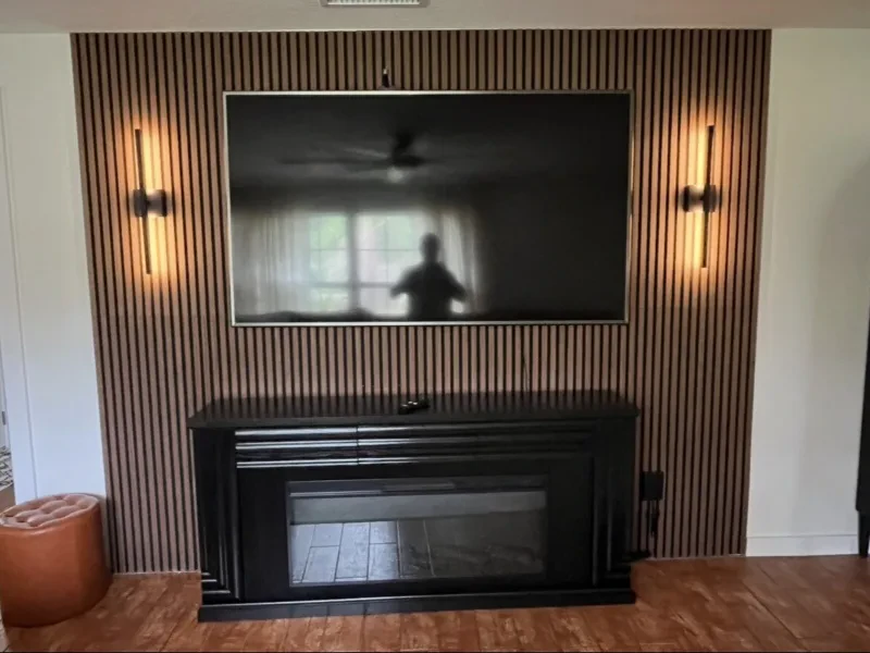 A wall of vertical wood slats with a black fireplace, a television, and a lit sconce on each side.