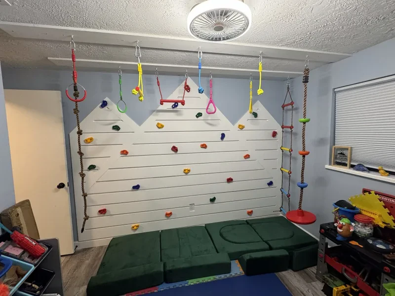 A white climbing wall shaped like three peaks, with colored holds, hanging rings, and a rope ladder.