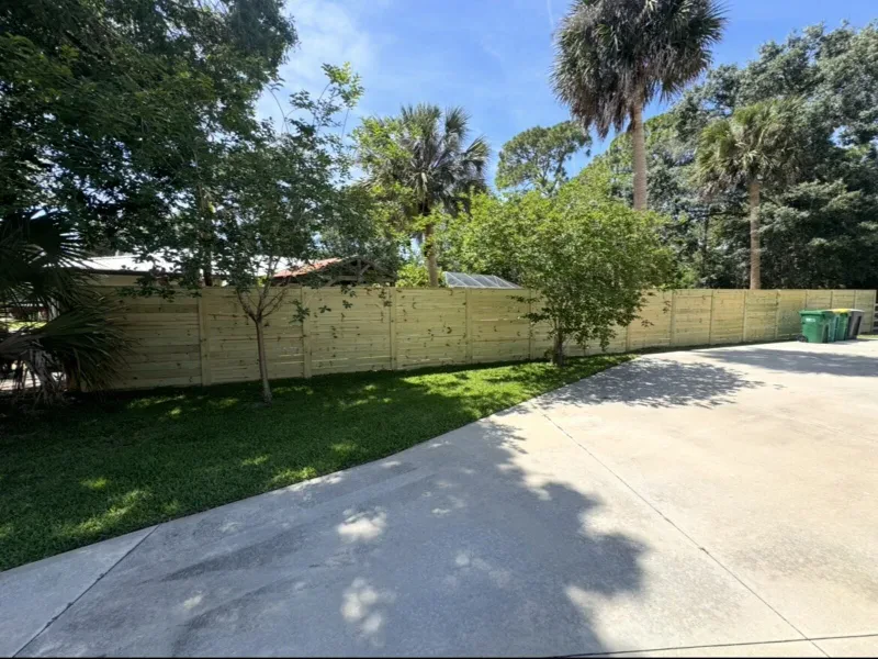 A long light-wood fence of horizontal slats, with grass and a driveway in front and palms behind.