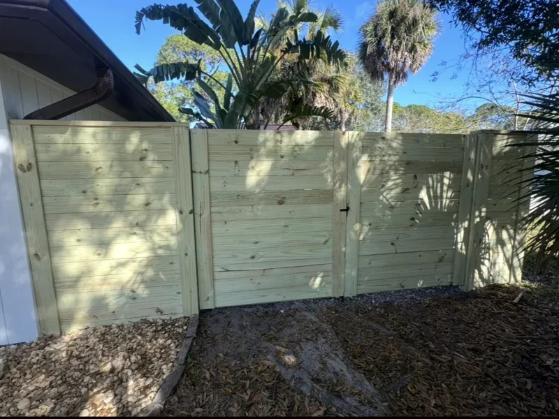 A light-wood gate of horizontal slats in a matching fence, with a house wall at the left and palms above.