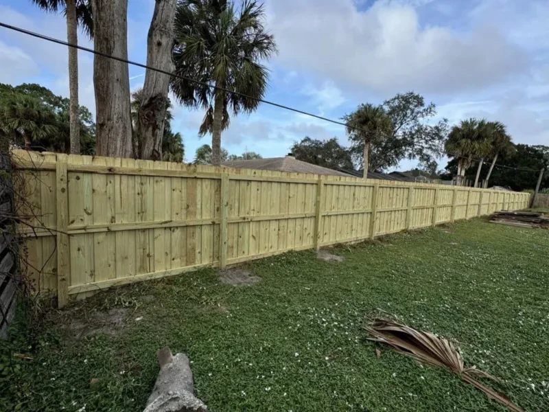 A light-wood horizontal-slat fence beside a grassy yard, with palm trunks and a roof behind it.