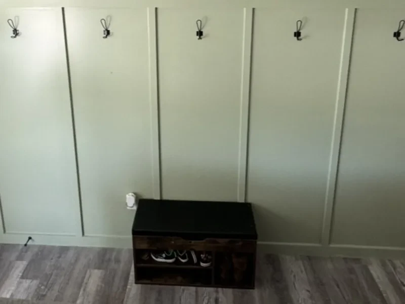 A sage green board-and-batten wall with black coat hooks and a dark shoe bench.