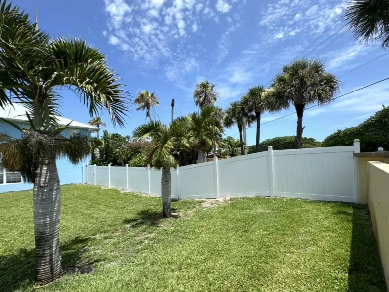 A white vinyl fence in a grassy yard, with palms and a blue house beside it.
