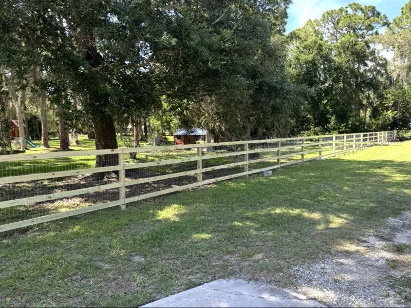 A light-wood three-rail fence with wire mesh, running through a grassy yard under oaks.