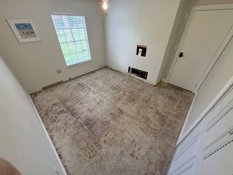 An empty room stripped down to bare plywood subfloor, wall cutout exposed in the corner.