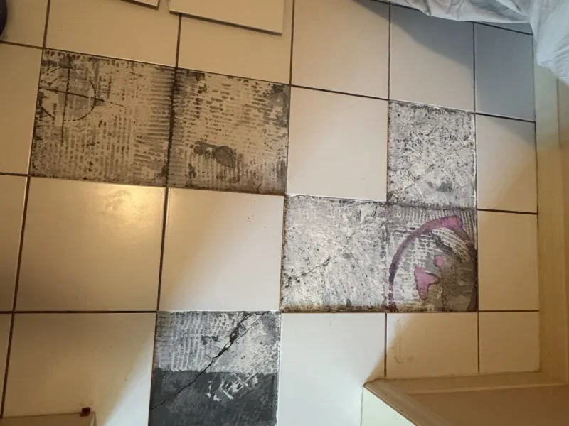 White floor tiles with several damaged tiles marked in dark patches, and a cabinet edge at the lower right.